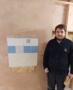 A young man in work clothes, standing next to a wall he has just plastered and tiled