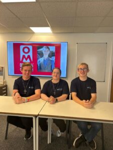 Three young people seated at a table and facing the camera. They are wearing Oliver McGowan's Campaign t-shirts and have a picture of Oliver on a large TV screen behind them.