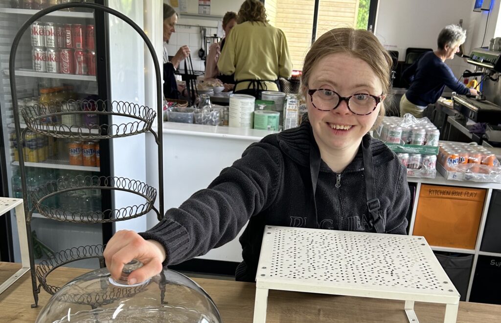 A young lady taking part in work-related learning at a cafe. She is standing behind the cafe's serving counter, smiling at the camera
