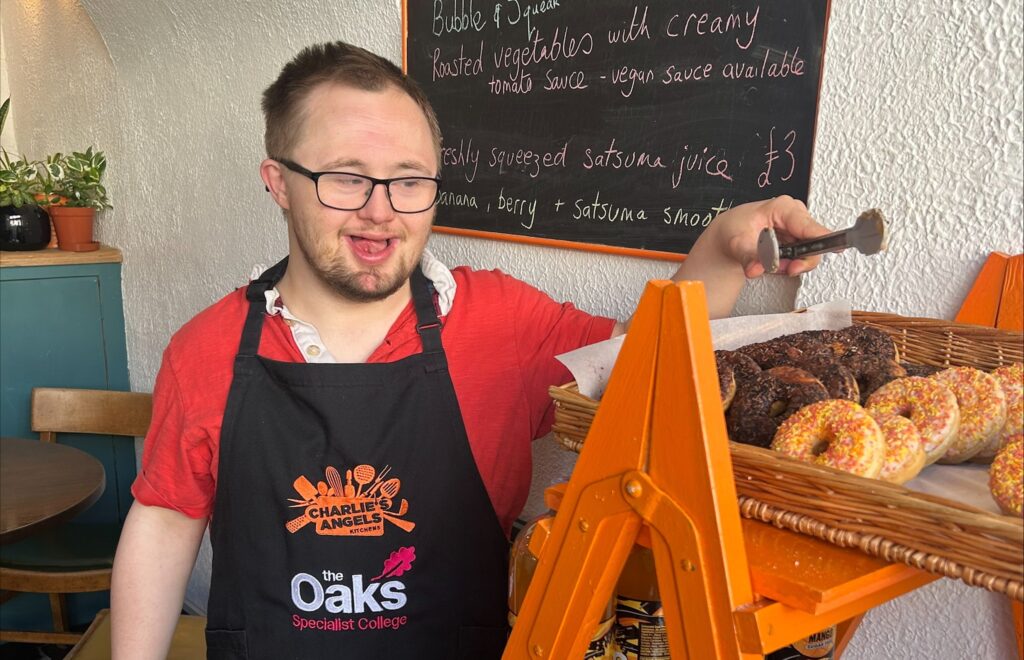 A young man in a serving apron picks up a doughnut with a set of steel tongs
