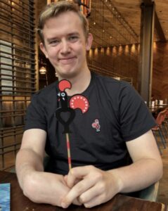 A young man in a Nando's staff t-shirt, seated at a table and smiling at the camera