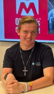 A young man seated at a training room table, smiling for the camera