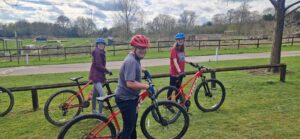 Three young people on mountain bikes in a green field with wooden fencing